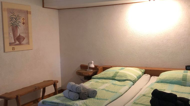 Ein gemütliches Schlafzimmer mit einem Doppelbett und modernen Bettwaren. Eine Holzbank, eine Deckenleuchte mit integriertem Ventilator und ein Kunstwerk ergänzen die Einrichtung.
