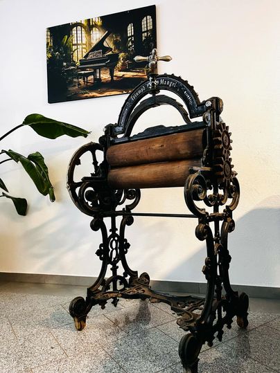 Een oude drukpers van metaal en hout staat in een kamer. Op de achtergrond is een afbeelding van een piano aan de muur te zien.