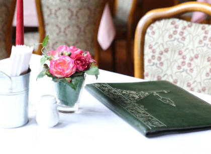 Een elegante tafel in een restaurant met een groen menu en een vaas met roze bloemen. Op de achtergrond zijn versierde stoelen te zien.
