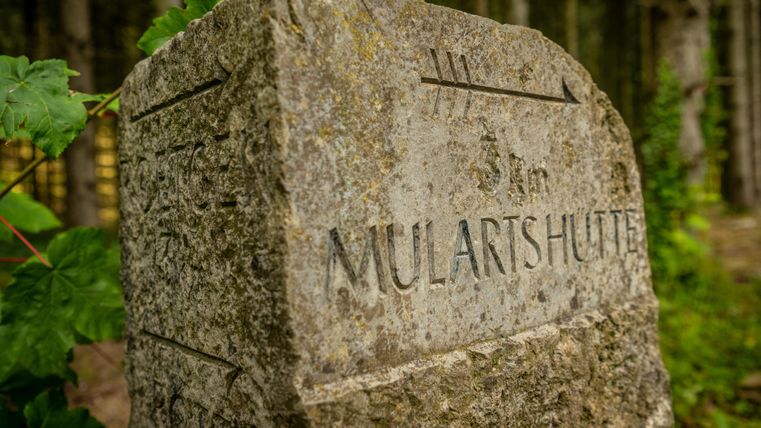 Un panneau en pierre avec l'inscription "MULARTHÜTTE". Entouré d'arbres et de verdure.