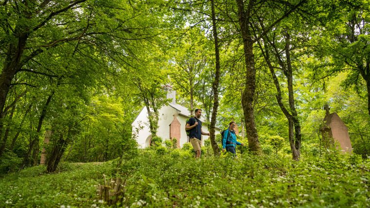 Twee wandelaars lopen door een bos met een klein wit kapelletje op de achtergrond.