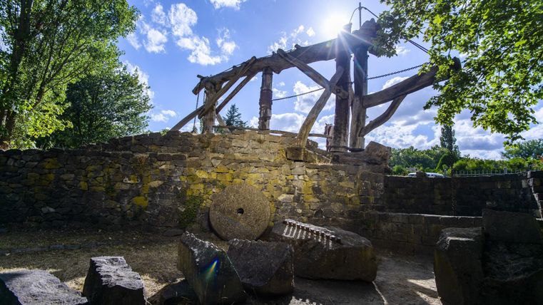 Ancien moulin en pierre avec construction en bois et meules en plein air, entouré d'arbres et d'un ciel bleu.