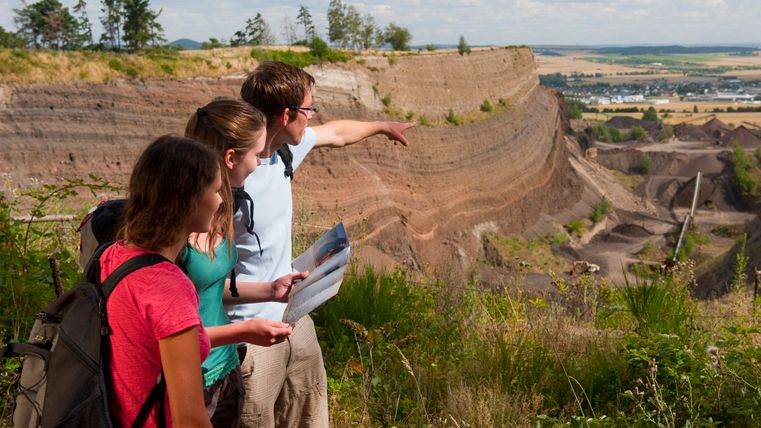Drei Personen stehen am Rand eines Steinbruchs und schauen in die Ferne. Eine Person zeigt mit dem Finger, während eine andere eine Karte hält.