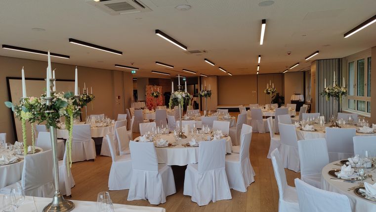 Een elegant versierde evenementenruimte met ronde tafels en witte tafellakens. De tafels zijn versierd met bloemstukjes en kandelaren.