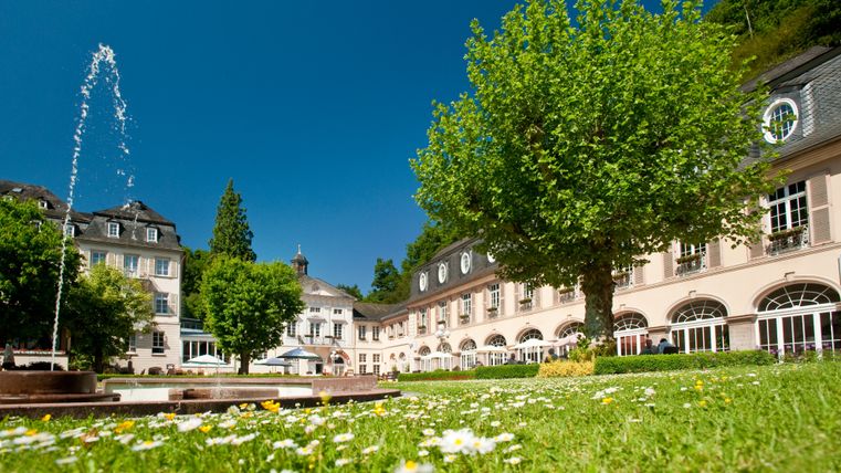 Ein gepflegter Kurgarten mit einem historischen Gebäude im Hintergrund und einem Brunnen im Vordergrund.