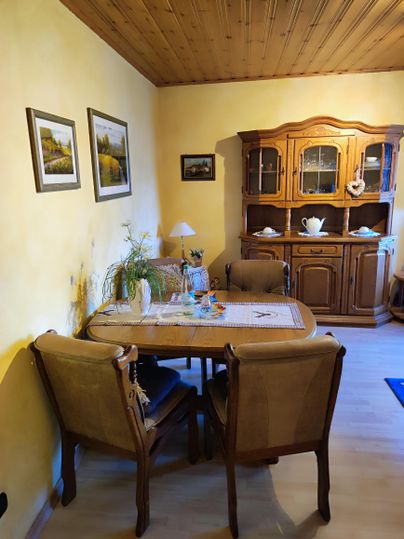 A cozy dining area with a wooden table and chairs. Pictures are hanging on the wall, and there is a cabinet with dishware.