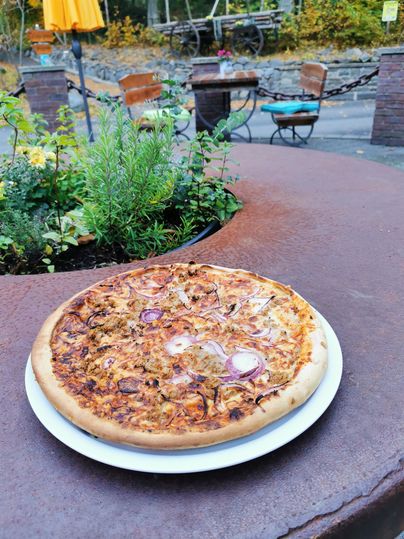 Une pizza fraîche sur une table rustique. En arrière-plan, on peut voir quelques sièges et des plantes.