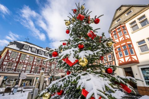Een versierde kerstboom bedekt met sneeuw staat voor vakwerkhuizen in Adenau, onder een blauwe hemel.