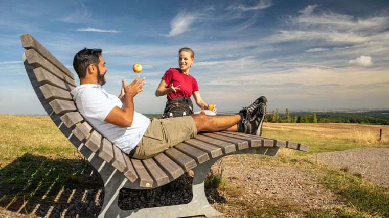 Two people on a curved bench in nature, one juggling with an apple.