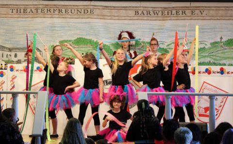 A group of children is performing a dance on a stage. They are wearing black T-shirts and colorful tulle skirts and are holding colorful sticks in their hands.