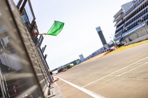Eine grüne Flagge wird an einer Rennstrecke geschwenkt. Im Hintergrund sind die Tribünen und eine Rennsäule sichtbar.