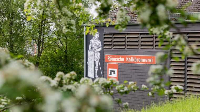 Gebouw met bord 'Römische Kalkbrennerei' en afbeelding van een Romeinse soldaat, omringd door bomen en bloemen.