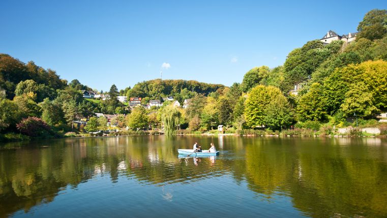 A pond in Blankenheim with a rowing boat and two people, surrounded by trees and houses on a hill.