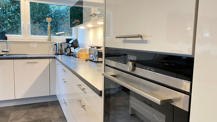A modern kitchen with white furniture and a black oven. Light comes in through large windows, and several kitchen appliances are visible.