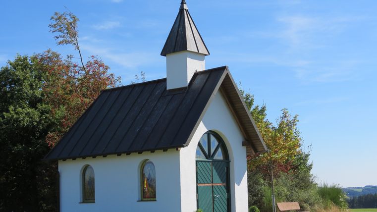 Kleine witte kapel met groene poort en kruis op het dak, omgeven door bomen en weiland.