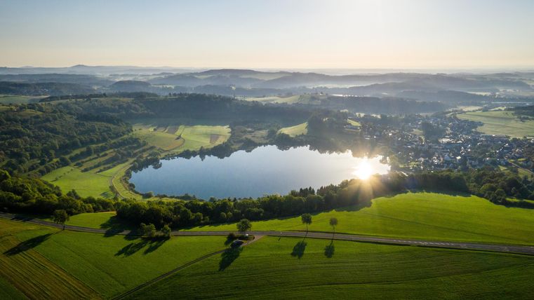 Eine weite Landschaft mit einem klaren See und grünen Feldern. Die Sonne scheint und sorgt für eine friedliche Atmosphäre.