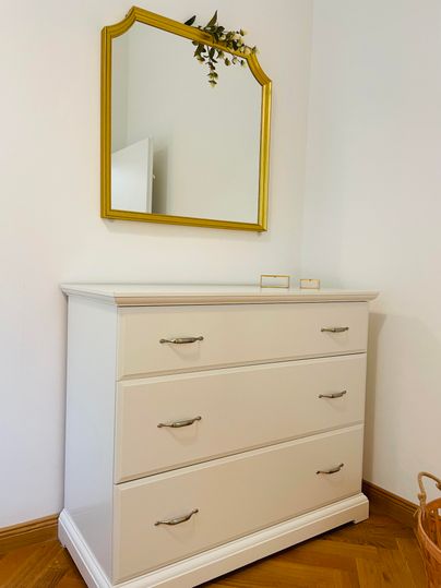 Une élégante commode avec trois tiroirs et un miroir doré au-dessus. La pièce a un sol en bois chaud et une atmosphère lumineuse.