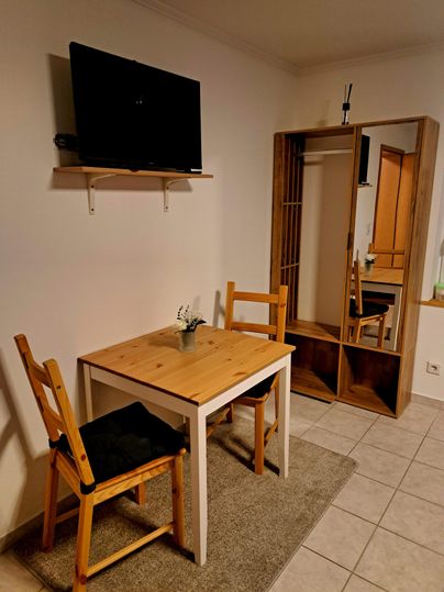 Houten tafel met twee stoelen in licht hout. Bovenaan is de televisie aan de muur bevestigd