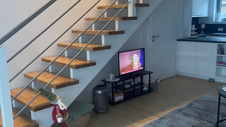 Eine moderne Treppe mit Holzstufen und einem eleganten Geländer. Im Hintergrund steht ein Fernseher auf einem niedrigen Tisch in einem stilvollen Wohnraum.