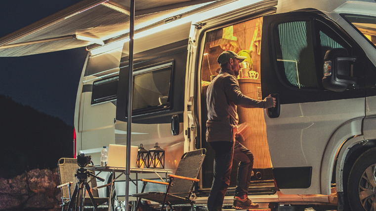 Une personne monte dans son camping-car. Dehors, il fait noir, la lumière à l'intérieur est allumée. Un pavillon est tendu sur le côté du camping-car.