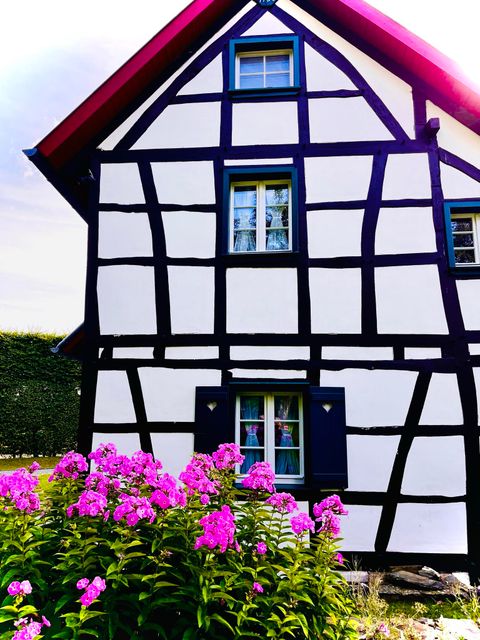 Une maison à colombages traditionnelle avec des murs blancs et des poutres noires. Devant la maison, des fleurs colorées fleurissent.