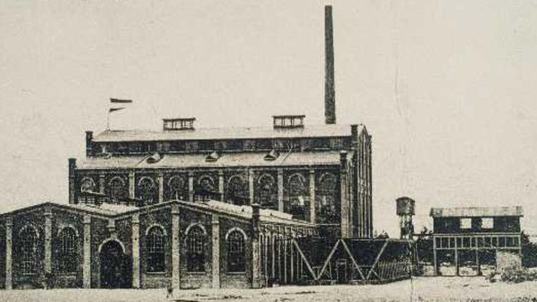 Eine historische Fabrikgebäude mit einem hohen Schornstein und großen Fenstern. Im Vordergrund sind weitere Gebäude und ein Wassertank sichtbar.