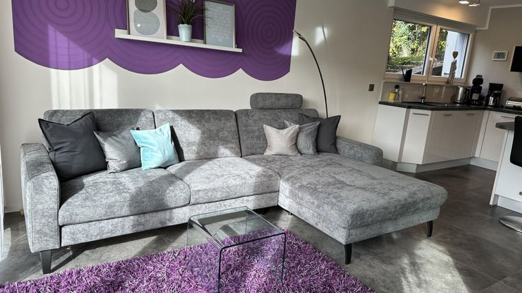 A modern living room with a gray sofa and colorful cushions. The room has a purple accent wall and a soft purple rug.
