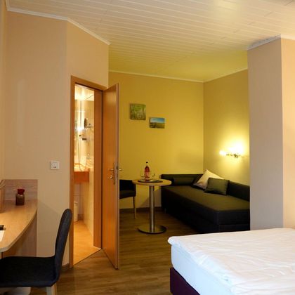 Een gezellige hotelkamer met een tweepersoonsbed, een tafel, een stoel en een zithoek. De muren zijn in zachte kleuren gehouden, en een televisie is aan de muur gemonteerd.