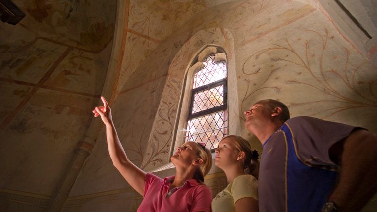 Drei Personen betrachten Fresken in einer Kirche mit einem gotischen Fenster.