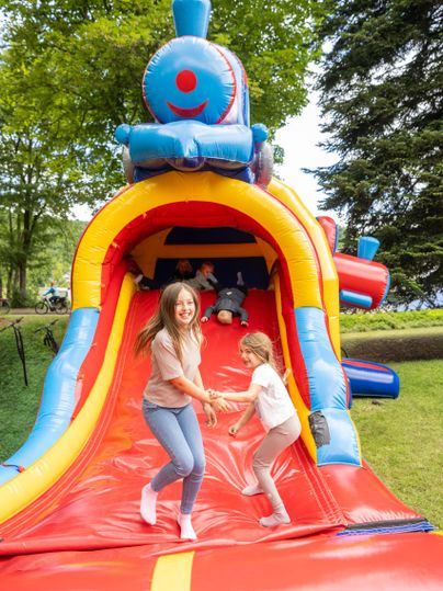 Een kleurrijk springkasteel in de vorm van een locomotief met blije kinderen die spelen. Op de achtergrond zijn bomen en een groen grasveld te zien.