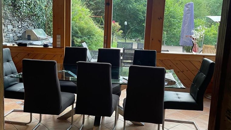 A modern dining area with a glass table and black chairs. Large windows offer a view of the green surroundings.