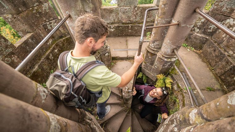 Twee personen klimmen een steile, stenen wenteltrap omhoog. Een persoon helpt de andere bij de beklimming in een oude toren.