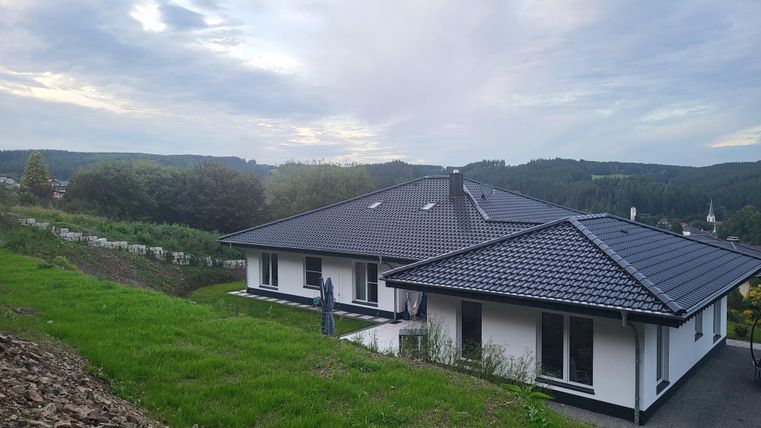 Een modern huis op een heuvel, omringd door groene weilanden. Op de achtergrond zie je een zacht heuvelachtig landschap onder een bewolkte hemel.