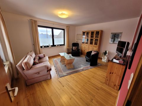 Ein helles Wohnzimmer mit einem gemütlichen Sofa und zwei Stühlen. Große Fenster bieten einen Blick nach draußen und der Boden ist aus Holz.