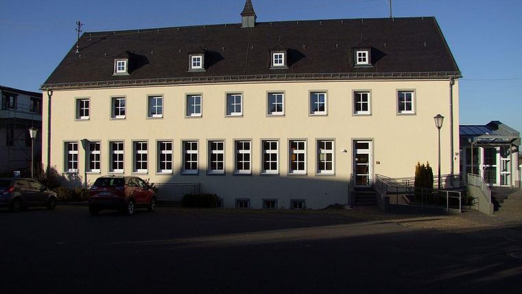 A large yellow building with several windows and a steep roof. It is situated in a quiet environment with a few parked cars.