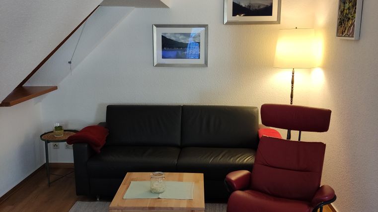 Ein gemütliches Wohnzimmer mit einem schwarzen Sofa und einem roten Sessel. Der Raum ist hell und einladend, mit Bildern an der Wand und einer Stehlampe.