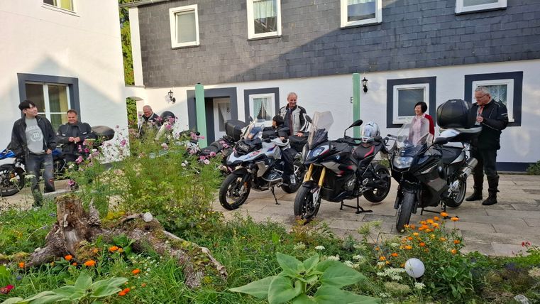 A group of motorcyclists stands in front of several motorcycles in a garden.