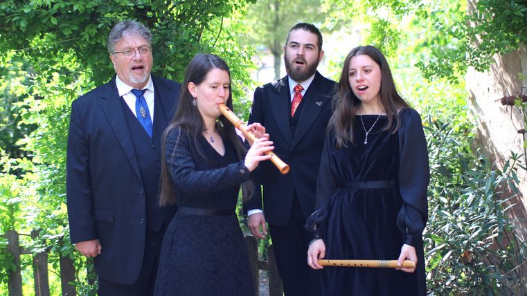 Eine Gruppe von vier Personen steht im Freien. Eine Frau spielt eine Flöte, während die anderen singen und musizieren.