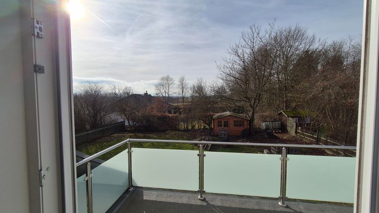 Vue d'un balcon sur un jardin avec des arbres et une petite cabane de jardin. Le ciel est clair et lumineux.