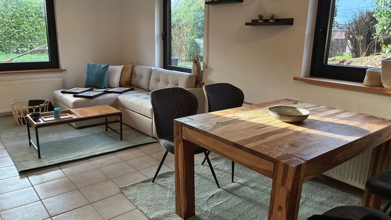 Een gezellige woonkamer met een moderne bank en een houten tafel. Grote ramen laten veel daglicht binnen en bieden uitzicht op de buitenlucht.