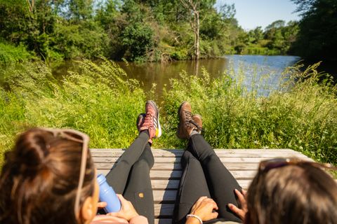 Zwei Personen entspannen auf einer Bank am Ufer eines Teiches, umgeben von grüner Natur.