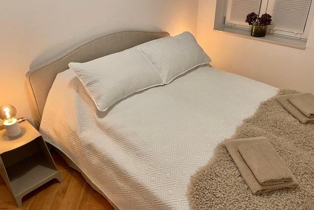 Ein gemütliches Schlafzimmer mit einem großen Bett und einer hellen, einladenden Atmosphäre. Neben dem Bett steht ein Nachttisch mit einer Kerze und frischen Blumen.