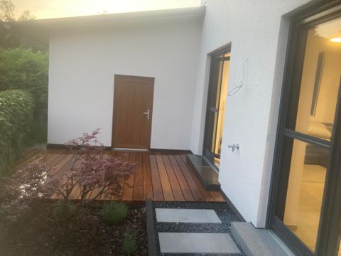 Eine moderne Hausansicht mit einem kleinen Holzdeck und einer Eingangstür. Im Vordergrund sind einige Pflanzen und der Weg aus Platten zu sehen.