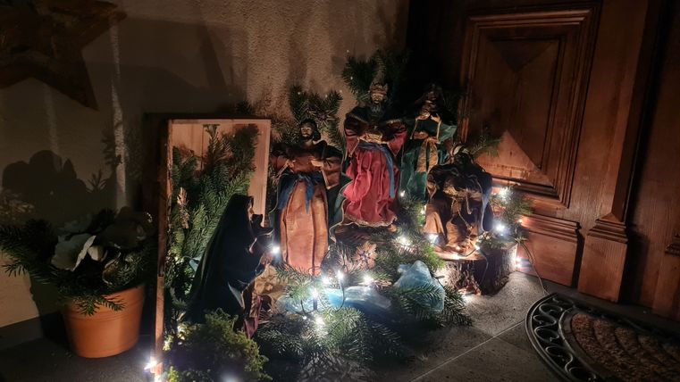 Weihnachtliche Krippenszene mit Figuren und Lichtern vor einer Holztür.