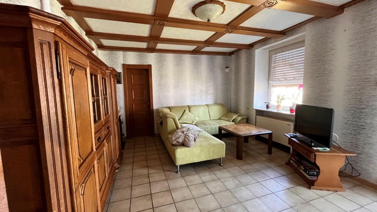 A cozy living room with a light green sofa and a wooden table. A television is on the wall, and a wooden cabinet is visible.