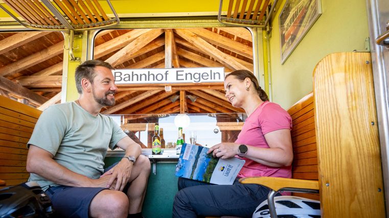 Ein Mann und eine Frau sitzen in einem Waggon und unterhalten sich. Im Hintergrund ist das Schild „Bahnhof Engeln“ sichtbar.
