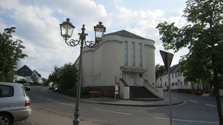Een rustige straat met een kerk en een lantaarn. Omringd door bomen en woongebouwen.