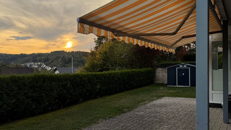 A garden with a sunshade and beautiful evening atmosphere. In the background, the setting sun and gentle hills are visible.