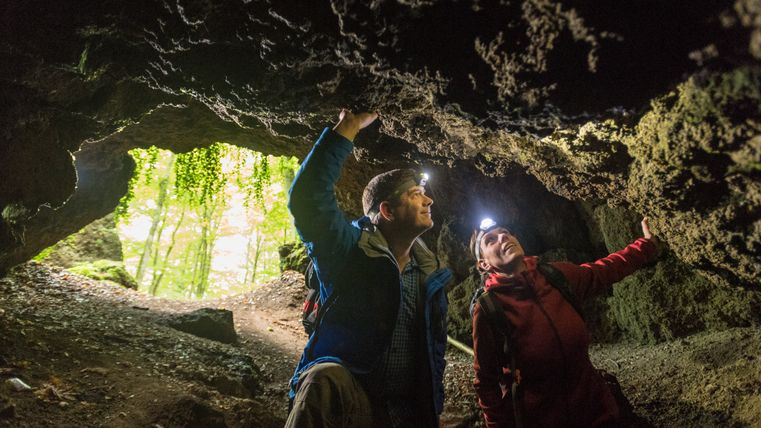 Deux personnes munies de lampes frontales explorent une grotte entourée de rochers et d'une forêt en arrière-plan.