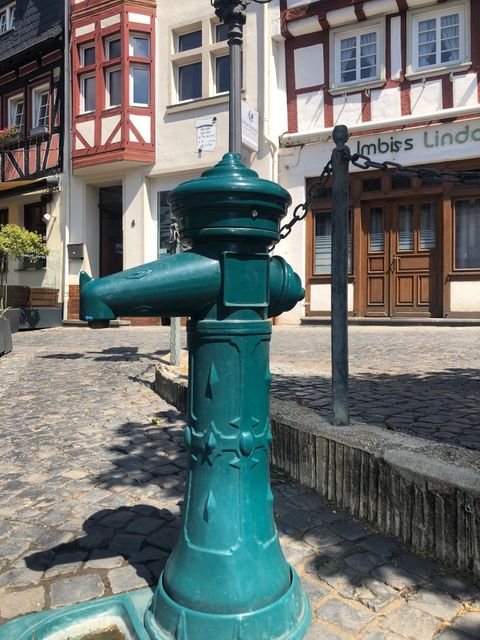 Un hydrant vert se trouve sur une place pavée. En arrière-plan, on peut voir des maisons à colombages traditionnelles et une petite restauration.